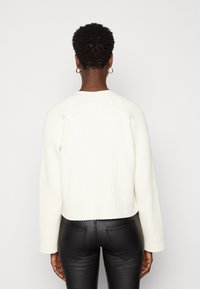 Vit ribbad kort tröja med långa ärmar, rund halsringning och textilstruktur, kombinerad med åtsittande svarta byxor.