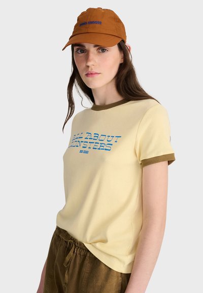 Femme portant un t-shirt beige avec des inscriptions bleues, des bordures marron, un pantalon marron avec cordon de serrage et une casquette marron, tournée légèrement vers la gauche sur fond blanc.