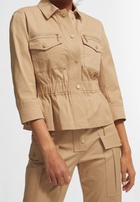Chaqueta utilitaria beige con bolsillos con botones y cremalleras, combinada con pantalones cargo a juego con bolsillos de solapa y detalles de cremallera.