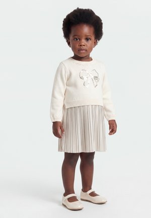 Bambino piccolo con capelli corti e ricci che indossa un maglione crema con un fiocco, gonna plissettata e scarpe Mary Jane crema, in piedi contro uno sfondo bianco.