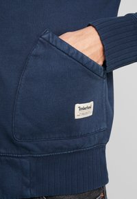 Sudadera azul marino con un gran bolsillo frontal, puños acanalados y una etiqueta de Timberland cosida en el bolsillo. Se puede ver la suave textura del tejido.