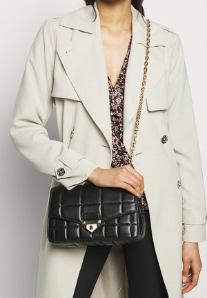 Zwarte doorgestikte handtas met een goudkleurige sluiting en kettingriem, vastgehouden in een hand. Heeft een rechthoekige vorm en een gladde, getextureerde uitstraling.