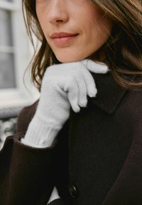 Fehér kötött kesztyű bordázott mandzsettával, egy barna kabáton pihenve. A kesztyűk puha textúrával és testhezálló kialakítással rendelkeznek.