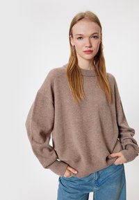 Młoda kobieta z długimi blond włosami, nosząca duże kolczyki w kształcie kół, luźny brązowy sweter z dzianiny i niebieskie dżinsy, stojąca z rękami w kieszeniach.