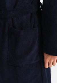 Mørkeblå frottébadekåpe med knytet midje, med en frontlomme og strukturert stoff. Mykt, absorberende materiale med lange ermer.