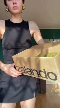 Henkilö, joka wearing mustan hihattoman mekon, pitää avointa pahvilaatikkoa Zalando, jonka sisällä on vihreä tuote, seisoo auringonvaloisassa huoneessa.