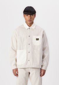 Striped beige jacket with a collar, two front pockets, and button closure. Fabric has a soft texture, with a small brand patch on the chest.