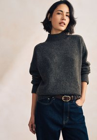 Pull en maille gris foncé avec col montant, poignets retroussés et coupe décontractée, associé à un jean en denim foncé et une ceinture marron avec boucle dorée.