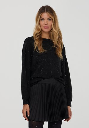 Donna con lunghi capelli biondi che indossa un maglione a pois neri, una gonna plissettata nera e calze a pois coordinate su uno sfondo neutro.