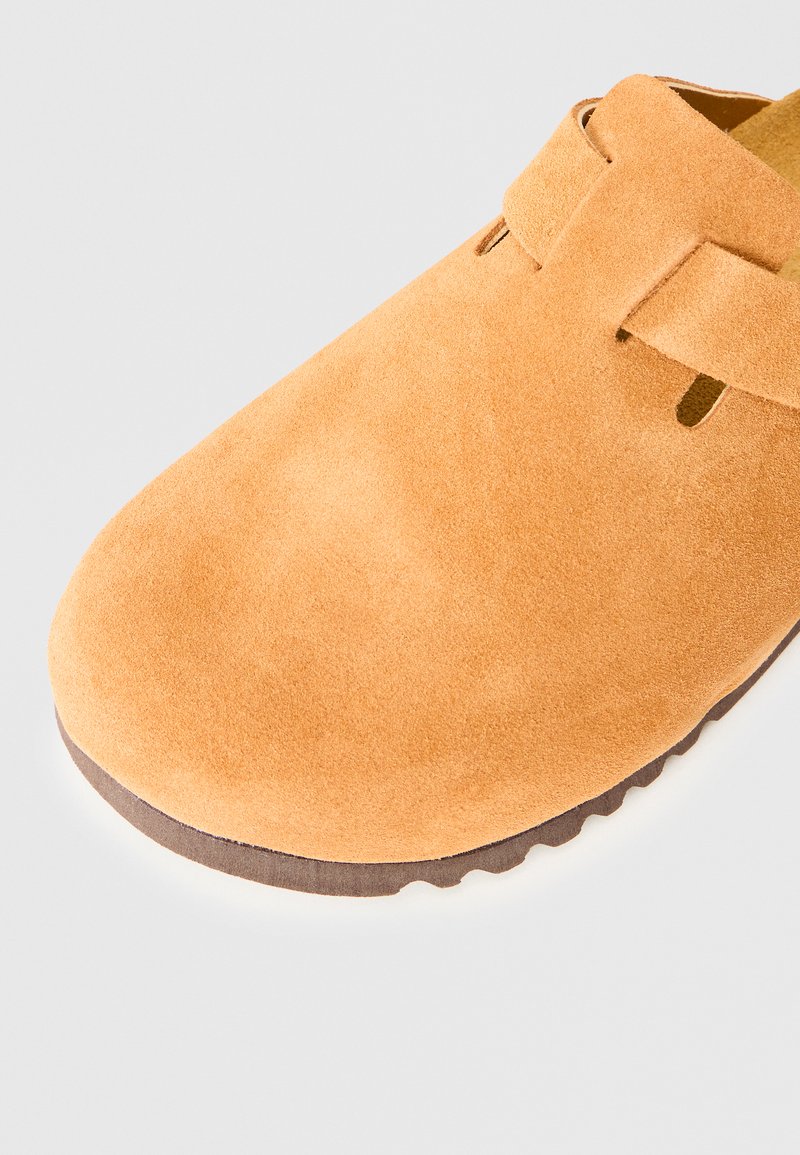 Hellbraune Wildlederschuhe mit Riemendetail und einer geriffelten dunkelbraunen Sohle auf einem hellen Hintergrund.
