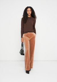 Camisa de manga larga marrón con canalé y cuello, combinada con pantalones de pana anchos color marrón claro. Los accesorios incluyen un pequeño bolso negro.