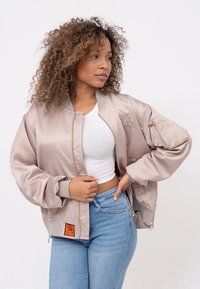 Giacca bomber rosa chiaro in satin, con chiusura a zip, polsini a coste e un'etichetta arancione sull'orlo. Il modello indossa una maglietta bianca e jeans.