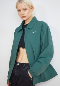 Zöld, könnyű dzseki lapos gallérral, patent gombokkal és fehér Nike logóval. Fekete crop top és fekete farmernadrág felett viselve.