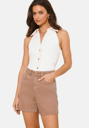 Kobieta ubrana w białą, bezrękawną bluzkę na guziki z kołnierzykiem oraz jasnobrązowe jeansowe szorty z wysokim stanem, pozująca z jedną ręką w kieszeni.