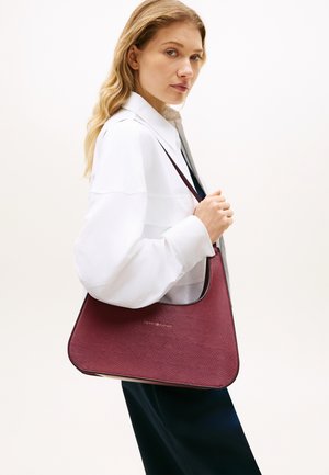 Βουργουνδί δερμάτινη τσάντα ώμου με υφή στην επιφάνεια, στρογγυλεμένο σχήμα και αντίθετο περίγραμμα. Διαθέτει χρυσό λογότυπο στο μπροστινό μέρος.