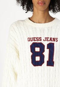 Maglione bianco a treccia con ricamo in blu e rosso della scritta "GUESS JEANS" e il numero "81". Collo a costine e dettagli di texture.