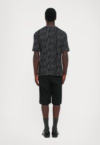 Man wearing black EA7 patterned T-shirt, black knee-length shorts, dark socks, and black shoes standing facing away on plain background.