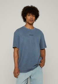 Jauns vīrietis ar cirtainiem matiem, ģērbies zila "ZAUDĒTA DZIMTENES" T-kreklā un gaiši zilos džinsos, stāv pretī vienotai fona.