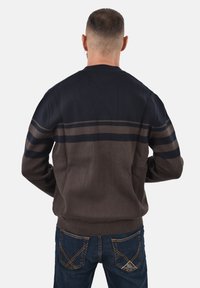 Uomo che indossa un maglione a righe blu navy e marrone e jeans blu scuro, in piedi di spalle contro uno sfondo chiaro e uniforme.