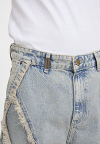 Jeans bleu clair délavés à l'acide avec un détail effiloché sur la couture latérale, portés avec un débardeur blanc côtelé, montrant la taille avant et l'étiquette de passants de ceinture.