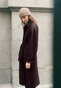 Mørkebrun uldfrakke med bælte, stort krave og knapmanchetter; parret med en lysebrun strikket hue. Tekstureret, varm stof.