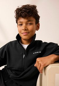 Schwarze Fleece-Zipjacke mit hohem Kragen und weißem Logo, über einem weißen Hemd getragen, mit glatter Textur und entspannter Passform.