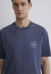 Navy blue crew-neck T-shirt made of cotton blend, featuring a circular white text graphic on the left chest area. Smooth texture.