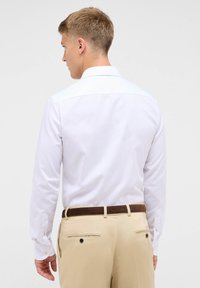Witte langemouwen shirt met knoopmanchetten en een stijve kraag, gecombineerd met tan broek met een bruine riem en achterzakken.
