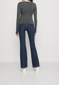 Langärmliges, enganliegendes graues Oberteil, kombiniert mit dunkelblauen, ausgestellt geschnittenen Jeans. Die Jeans haben Gesäßtaschen und einen sauberen Abschluss am Saum. Weiße Plateauschuhe.