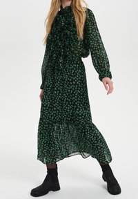 Lång, grön blommig klänning i genomskinligt tyg, med en volangkrage och ärmar, tillsammans med svarta ankelstövlar. Blommönster överallt.