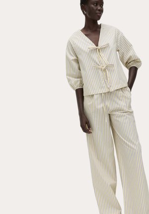 Donna con indosso un top a righe color crema a maniche lunghe con fiocchi e pantaloni a gamba larga a righe coordinati, in piedi contro uno sfondo semplice.