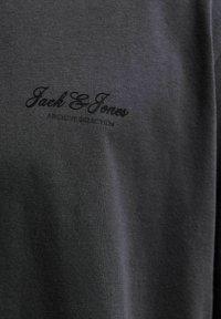 Σκούρο γκρι ύφασμα με το λογότυπο "Jack & Jones" και το κείμενο "ARCHIVE PRODUCTION" με μικρά μαύρα γράμματα τυπωμένο στο άνω αριστερό μέρος του στήθους.