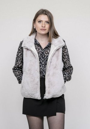 Jeune femme portant un gilet en fausse fourrure gris clair par-dessus un chemisier imprimé léopard noir et blanc et une jupe noire, debout devant un fond uni.