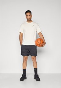 Mand i hvid Nike T-shirt, sorte Nike shorts og sorte sneakers, der holder en orange basketball mod en simpel lys baggrund.