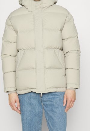 Giacca puffa beige chiaro con cappuccio, caratterizzata da un design trapuntato, grandi tasche frontali e polsini elastici. Il materiale appare morbido e isolato.