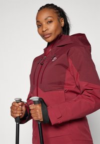 Veste imperméable bordeaux avec capuche zippée, poches avant et poignets ajustables. L'utilisateur tient des bâtons de trekking. Texture lisse, matériau durable.