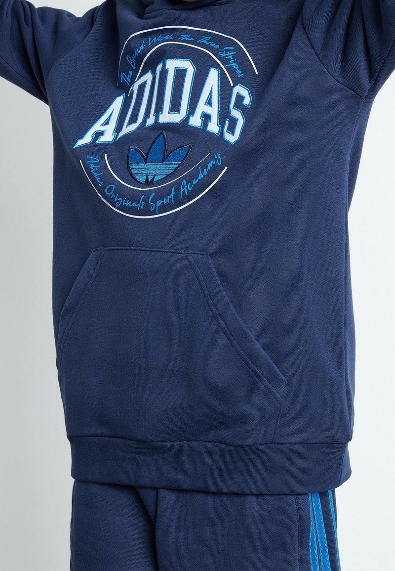 Sudadera Adidas de color azul marino con bolsillo frontal, gran logo y texto de Adidas en blanco y azul, combinada con pantalones marinos con rayas azules.