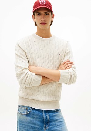 Joven con los brazos cruzados, vestido con un suéter de punto trenzado color crema, jeans azules y una gorra de béisbol roja con un emblema blanco.