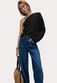 Un pull noir à une épaule associé à un jean bleu à taille haute à jambes larges et un sac en paille tressée avec des anses noires. Des bijoux en or complètent le look.