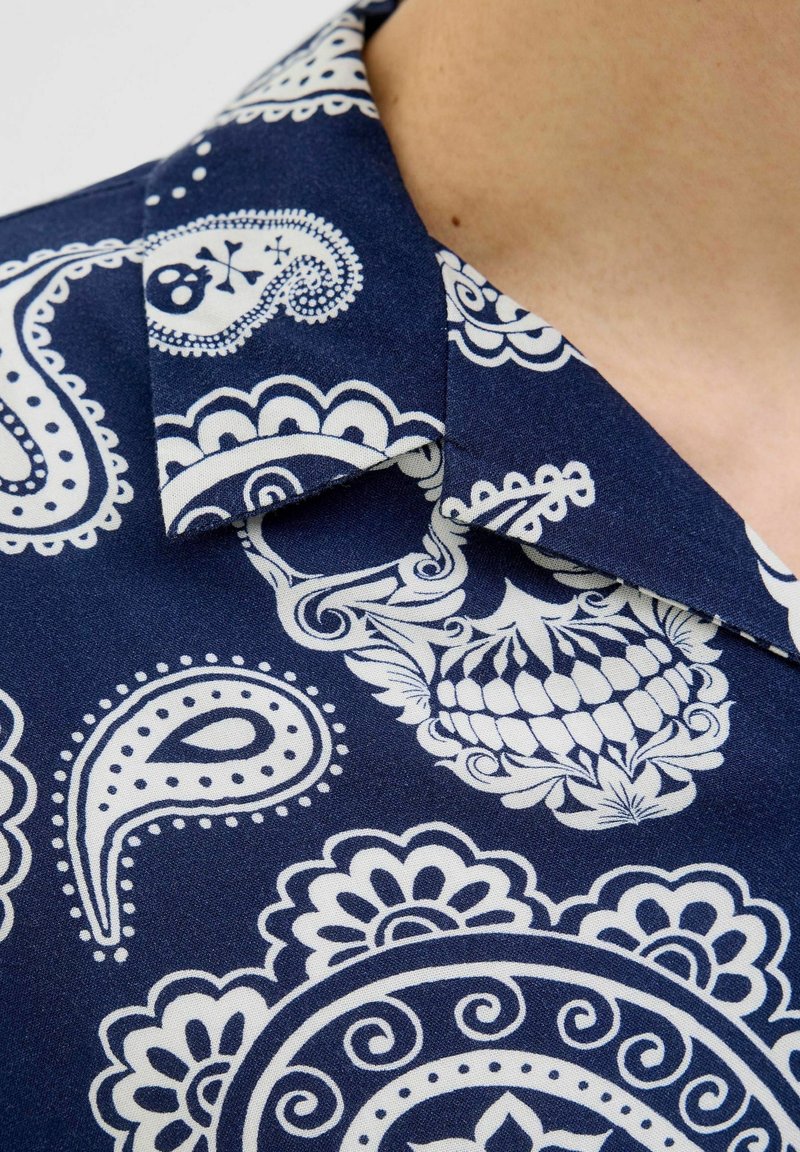Kortärmad skjorta med en marinblå bakgrund som har vita paisley- och blommönster; materialet är lätt bomull med krage.