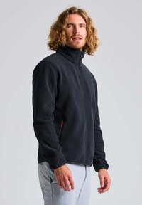 Schwarze Fleecejacke mit hohem Kragen und durchgehendem Reißverschluss, ausgestattet mit seitlichen Taschen und einer glatten Textur. Ideal zum Lagen-Look.