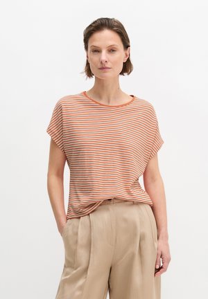 Woman with short brown hair wearing an orange and white striped short-sleeve shirt and beige high-waisted pleated pants, hands in pockets.