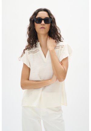 Blusa color crema con scollo a V, colletto arricciato e maniche corte a volant. Presenta dettagli in pizzo sulle spalle. Tessuto liscio.