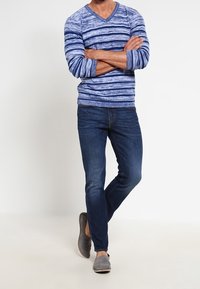 Blå- och vitrandig långärmad V-ringad tröja, kombinerad med mörkblå jeans och grå slip-on skor. Modellen står med armarna i kors.