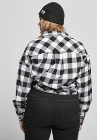 Donna di spalle che indossa una camicia a scacchi bianca e nera, pantaloni neri e un berretto di lana nero su uno sfondo semplice.