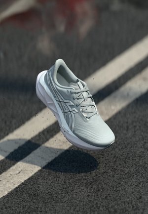 Zapatilla de correr gris claro con cordones en el aire sobre una carretera de asfalto con marcas blancas de carril y escombros negros dispersos.