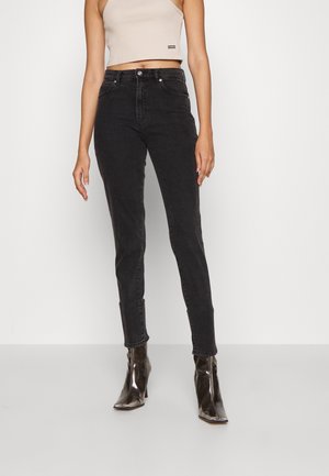 Kobieta ubrana w beżowy krótki top na ramiączkach, czarne obcisłe jeansy z wysokim stanem oraz błyszczące, czarne botki na obcasie z ostrym noskiem.