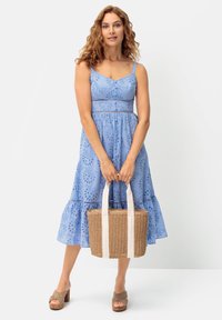 Svetlo modra obleka s dolžino do kolen, vezenimi cvetličnimi vzorci in trakovi za ramena. Model drži tkano torbico s kremnimi trakovi.