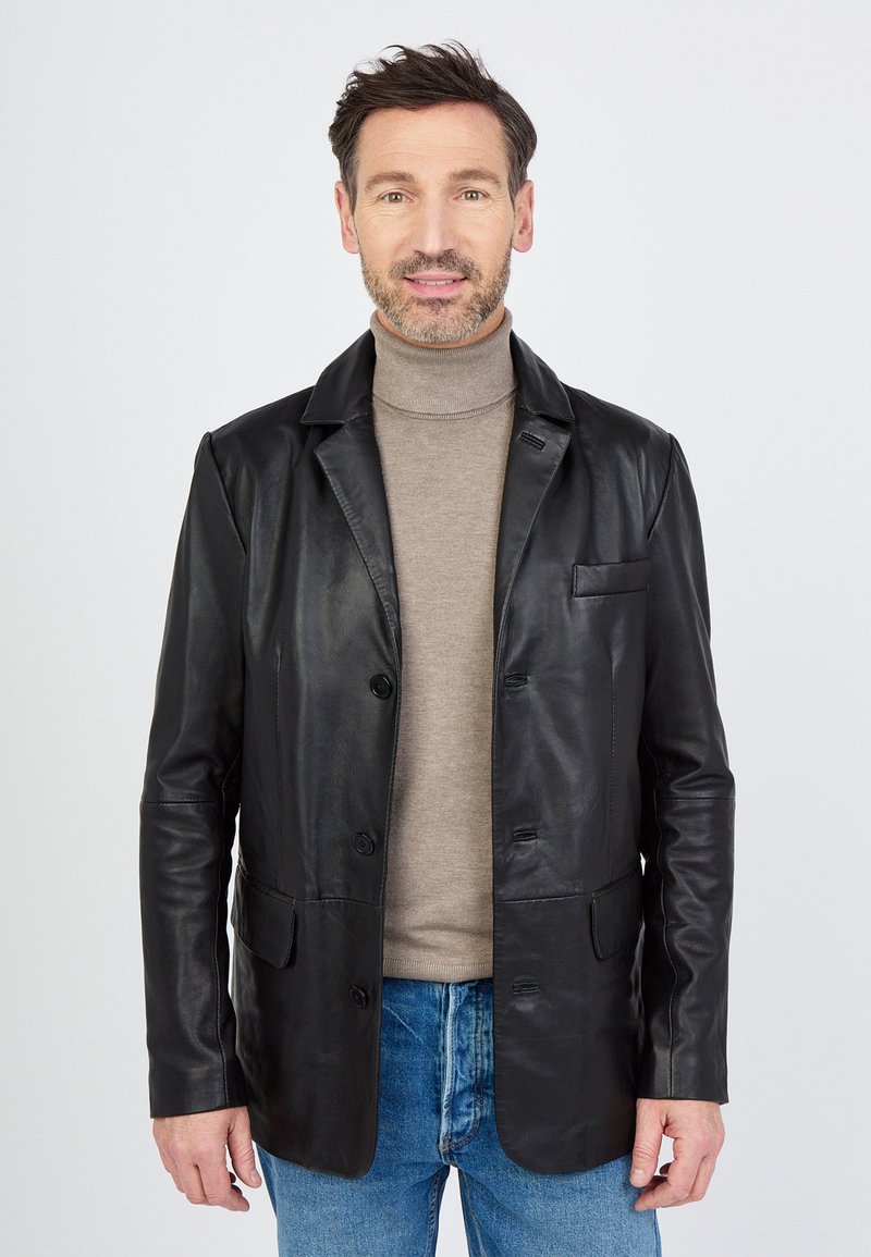 Czarna skórzana marynarka z dwoma przednimi kieszeniami i pojedynczym zapięciem na guzik, noszona na beżowym swetrze z golfem, w komplecie z niebieskimi dżinsami.