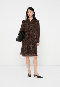 Sieviete ģērbjas brūnā leoparda raksta ceļa garuma kleitā, tur melnu kluču soma un valkā melnas smailes mūlītes.